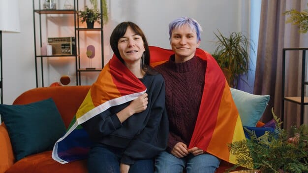 A close-up photo of two smiling women in China, holding hands and gazing affectionately at each other in a cozy living room that subtly includes rainbow-themed decor, symbolizing growing acceptance of LGBTQ+ relationships and their aspirations for family life.